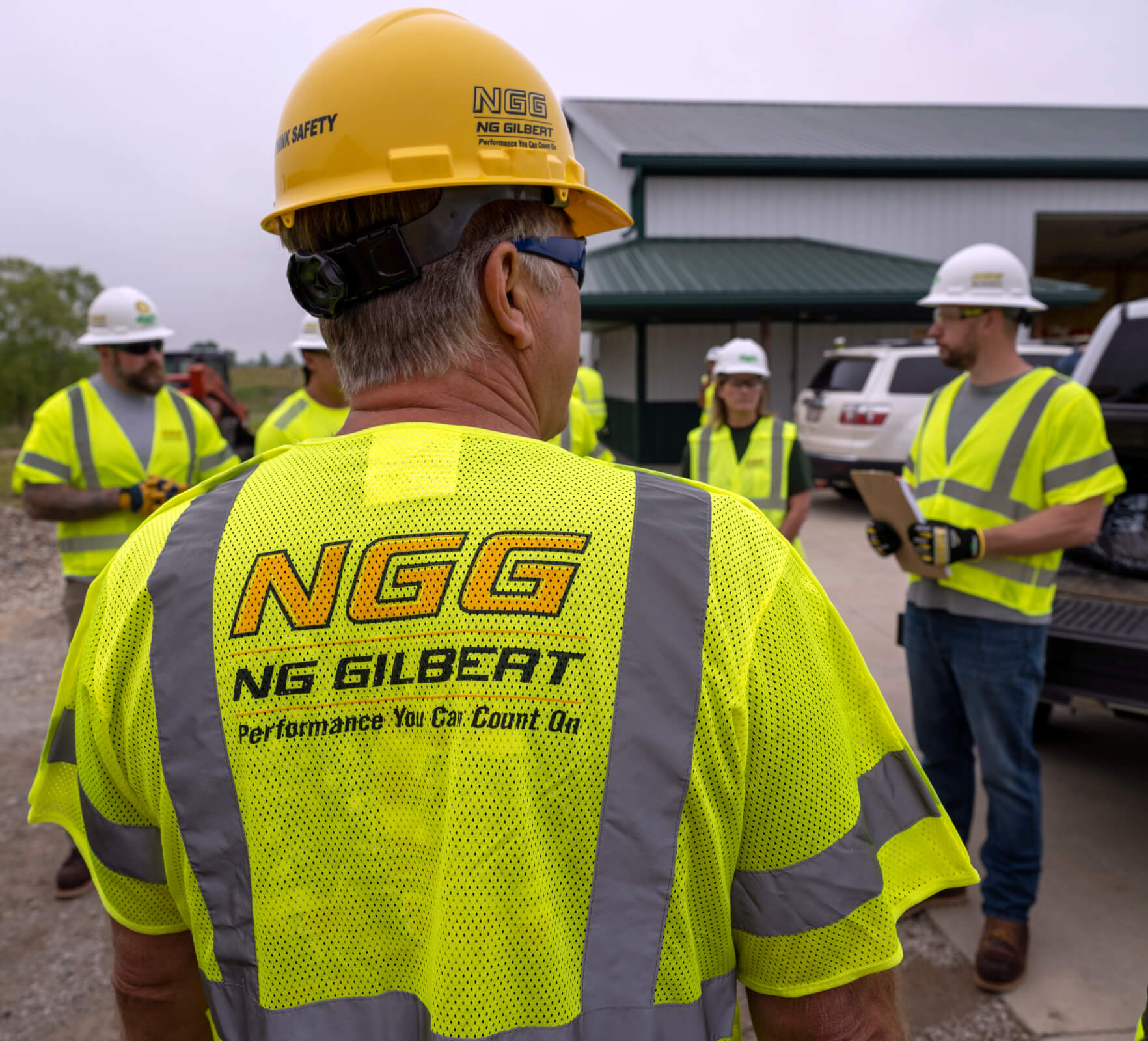 An NG Gilbert crew member with his back turned to the camera and facing other team members. All wear safety vests and hard hats as they stand outside a job site.