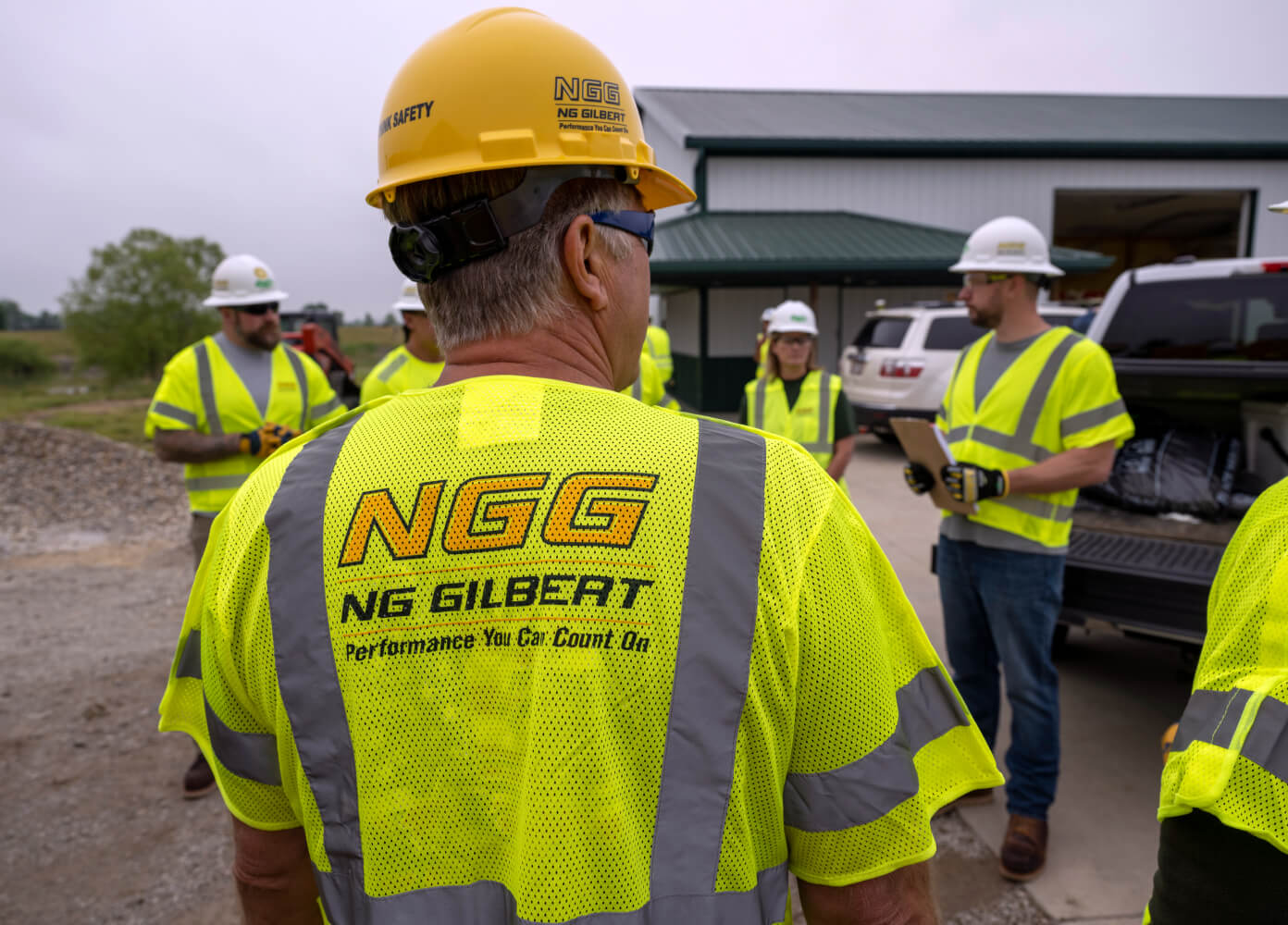 An NG Gilbert crew member with his back turned to the camera and facing other team members. All wear safety vests and hard hats as they stand outside a job site.