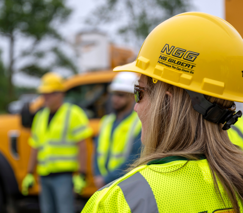 The back of the head of an NG Gilbert crew member. She wears a yellow hard hat, safety vest, and sunglasses as she looks on at other crew members.